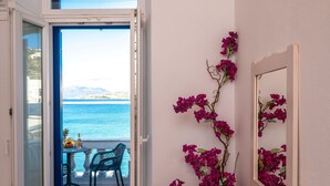 Een kamer met houten vloer, een tafel met stoelen, een bank, een deur die leidt naar een balkon met uitzicht op zee, en paarse bloemen aan de muur.