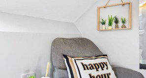 Un sillón gris con un cojín que dice 'happy hour', una planta pequeña y un cuadro con imágenes de plantas en la pared.