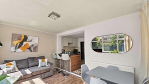 Un salon moderne avec un canapé gris, une table basse et un tableau au mur. On y trouve également un coin repas avec une table et des chaises, et une cuisine à l’arrière-plan.