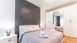 Een moderne slaapkamer met een behang, een groot bed met hoofdeinde, een nachtkastje met lamp, en een garderobe met spiegel.