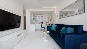 Ein modernes Wohnzimmer mit einem Flachbildfernseher, einem dunkelblauen Sofa mit türkisfarbenen Kissen und einem weißen Couchtisch. Im Hintergrund ist eine Küche zu sehen.
