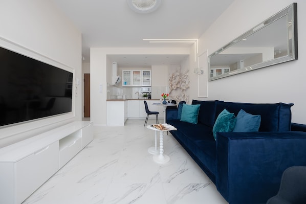 Un salon moderne avec un téléviseur à écran plat, un canapé bleu foncé agrémenté de coussins turquoise, une table basse blanche et, au fond, une cuisine.