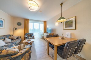Ein modernes Wohnzimmer mit einer Couch, einem Esstisch und Stühlen. Es gibt ein Fenster mit Vorhängen, eine Uhr an der Wand und eine Lampe.