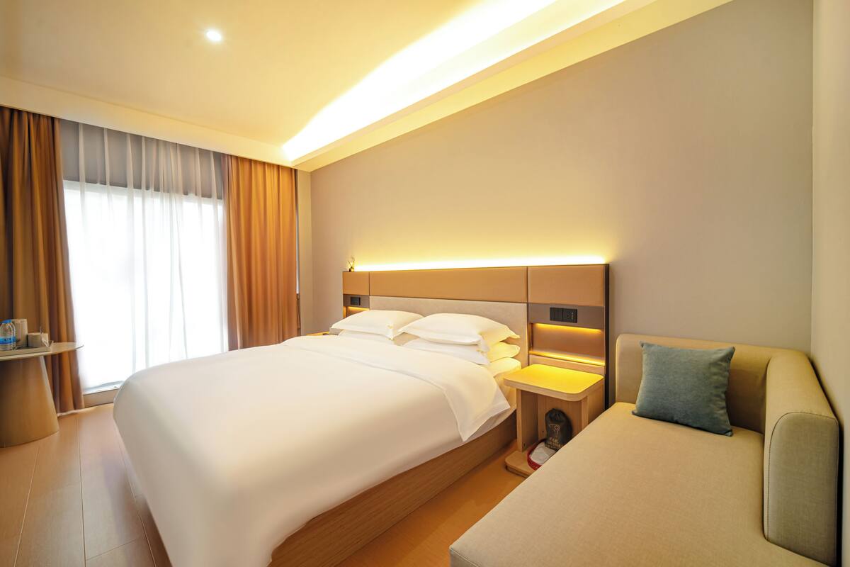 A hotel room with a large bed, bedside tables, a sofa, and a window with curtains.