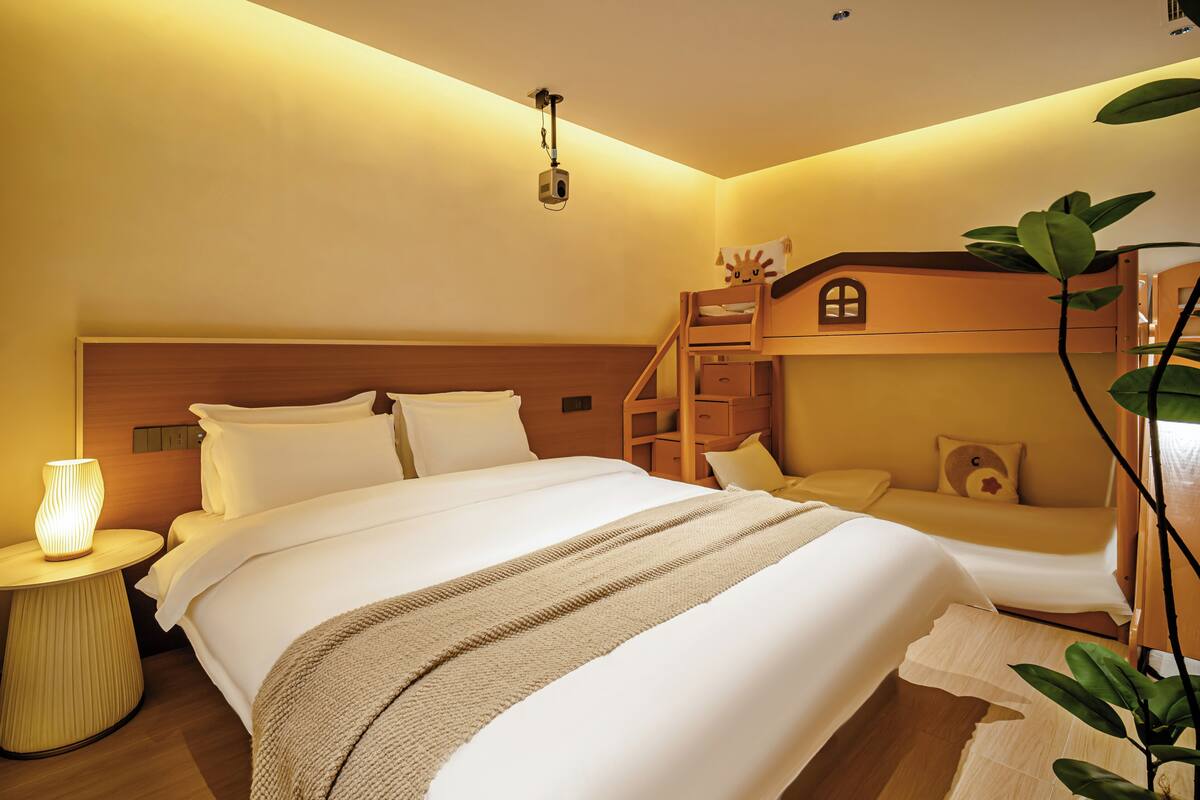 A hotel room with a large bed, a small desk, a nightstand, a wall-mounted projector, and a small wooden shelf with a ladder.