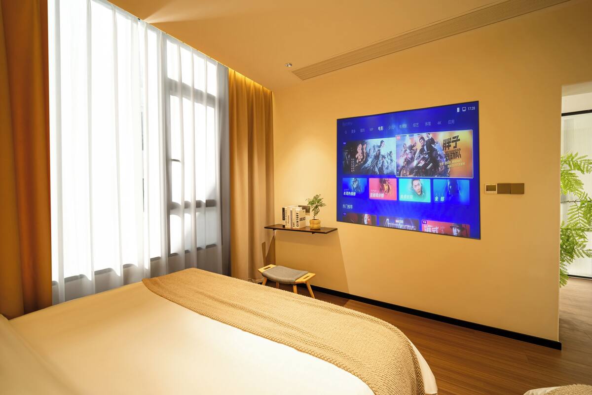 A hotel room with a large bed, a wall-mounted TV displaying a movie, a small table with a plant, and a window with curtains.