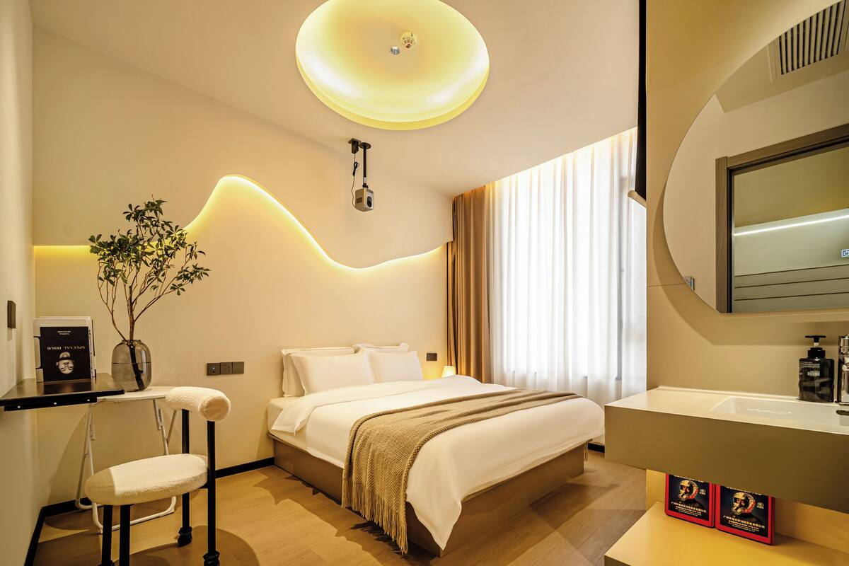 A modern hotel room with a bed, bedside table, chair, and a large mirror.