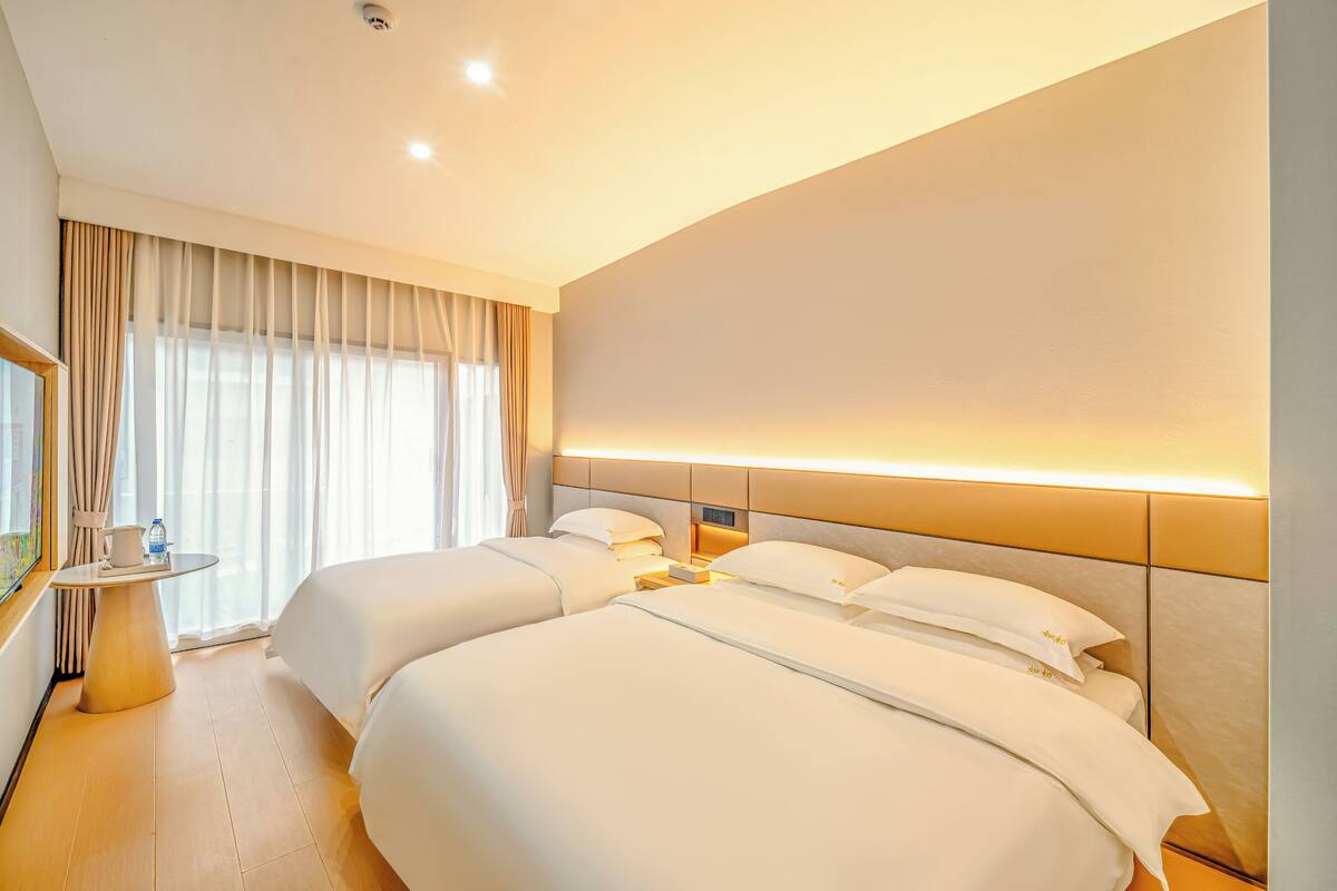 A hotel room with two beds, a bedside table with a lamp, a large window with curtains, and a wall-mounted light.