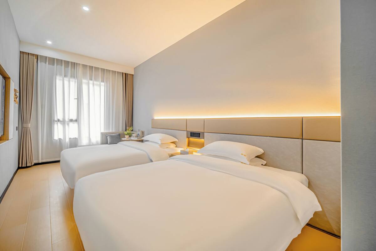 A hotel room with two beds, a bedside table, a lamp, and a large window with curtains.