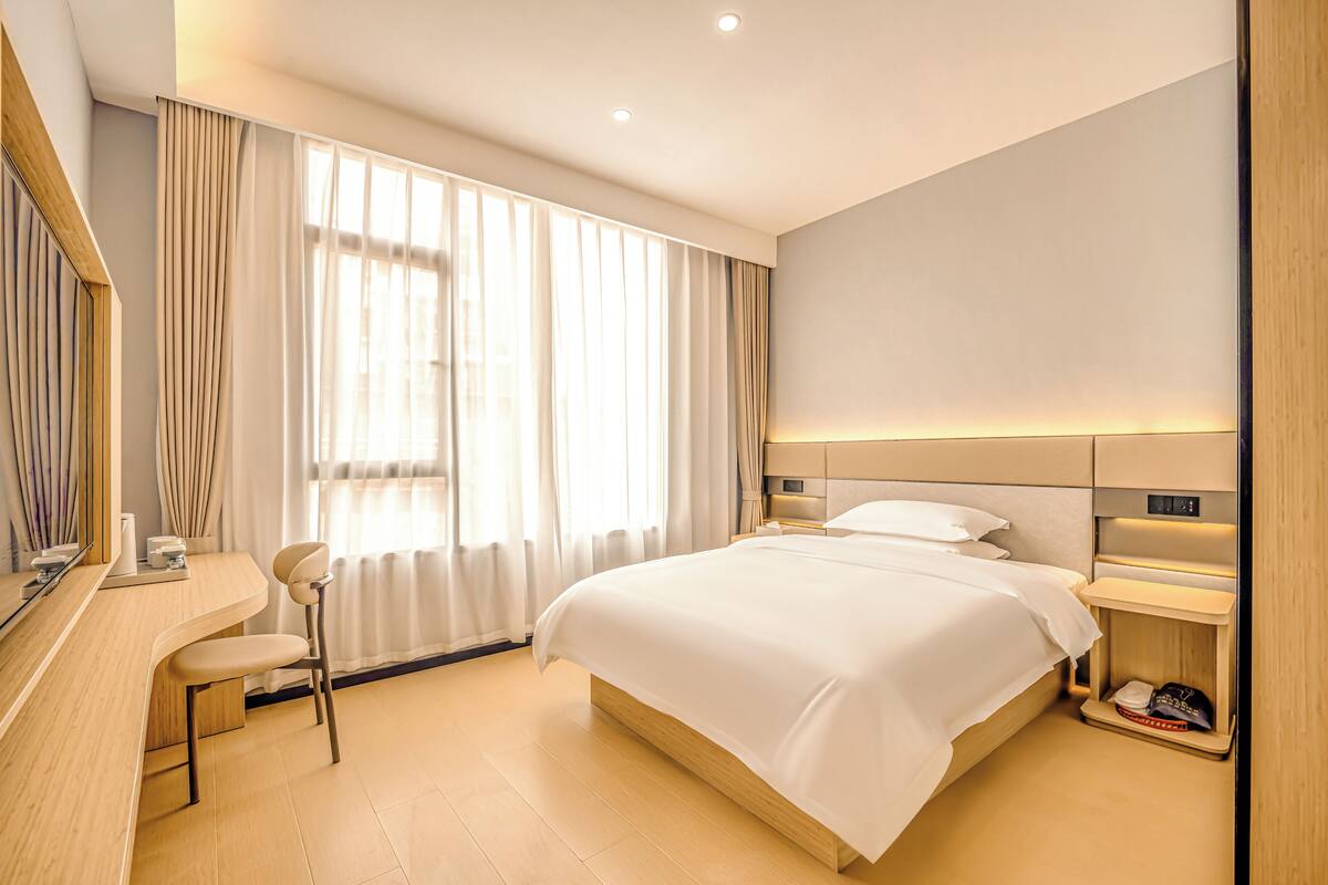 A modern hotel room with a large bed, a desk with a chair, a bedside table, and a mirror.