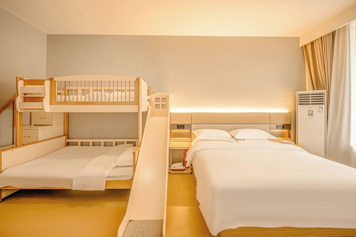 A hotel room with two beds, a bunk bed, a bedside table, a lamp, and a wardrobe.