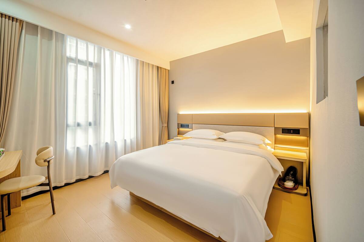 A hotel room with a bed, bedside table, chair, and a window with curtains.