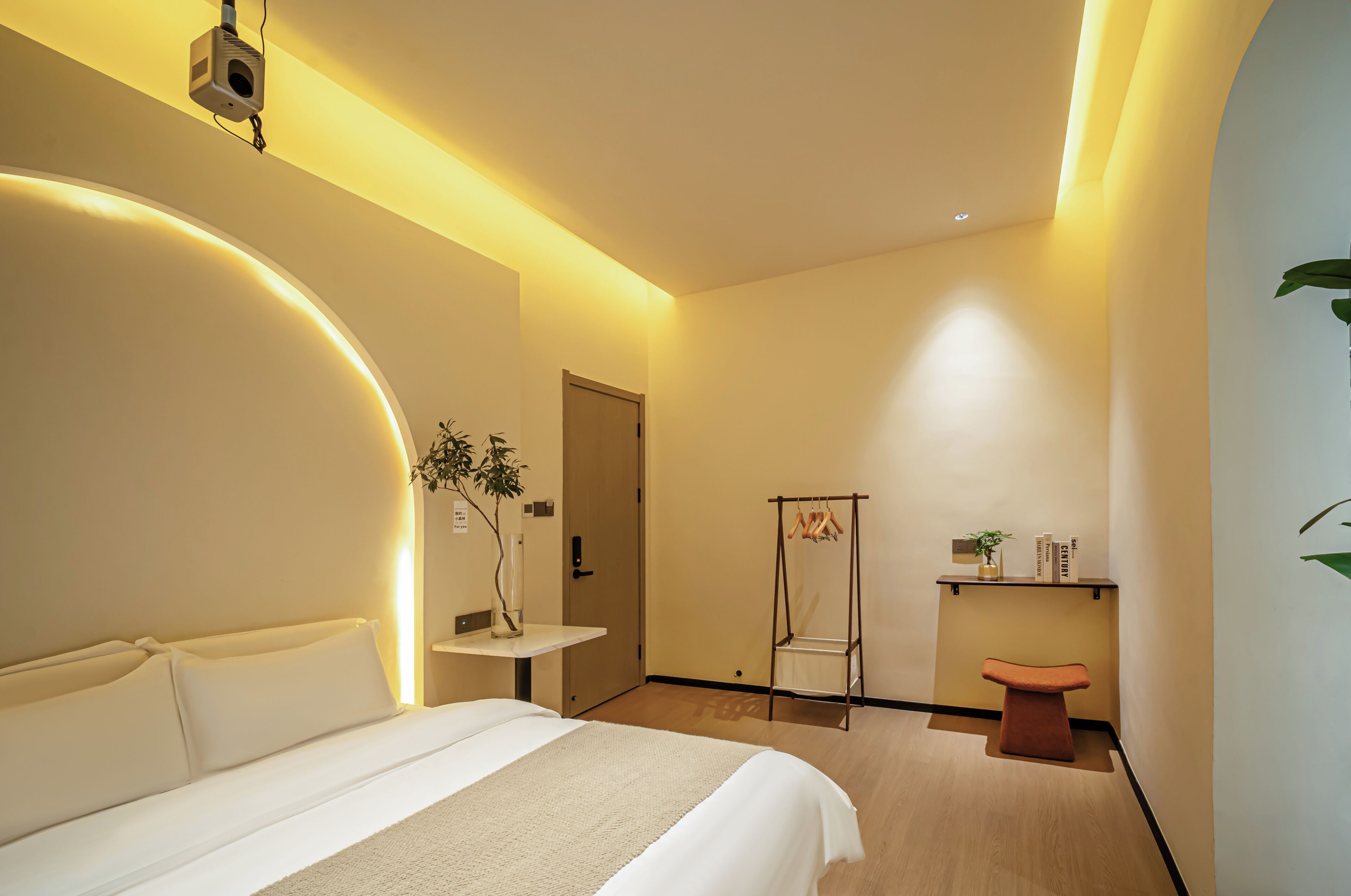 A hotel room with a bed, bedside table, a small desk, a chair, a shelf with books, and a plant.