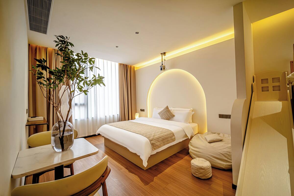 A hotel room with a bed, bedside table, chair, and a plant.