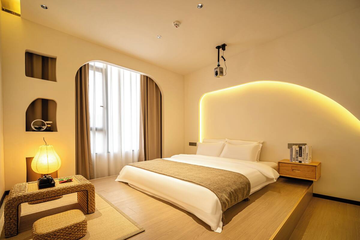 A modern hotel room with a bed, bedside table, lamp, and a view of the outdoors.