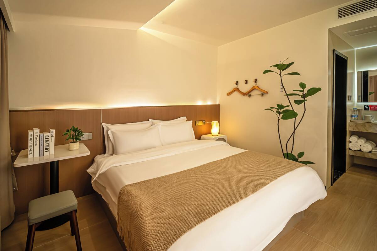 A hotel room with a bed, bedside table, a small stool, a wall-mounted plant, and a mirror.