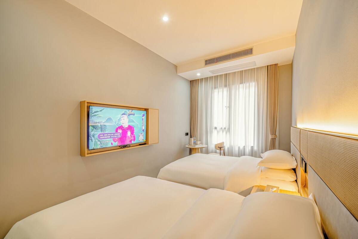 A hotel room with two beds, a television mounted on the wall, a small table, and a window with curtains.
