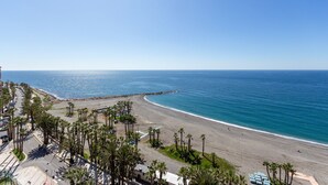 Uma praia com vista clara do oceano, uma estrada com carros estacionados e palmeiras.