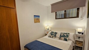 Une chambre à coucher avec un lit, des tables de chevet, une commode, une fenêtre avec des stores, une porte et un petit tableau accroché au mur.