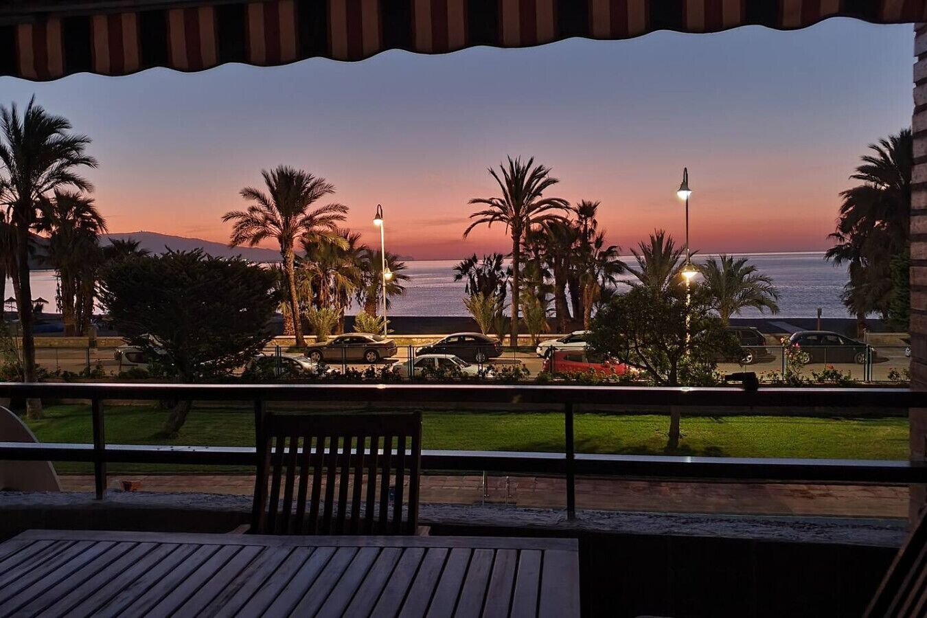 Une vue sur le coucher de soleil depuis un balcon, avec des palmiers, une piscine et une rue bordée de voitures garées.