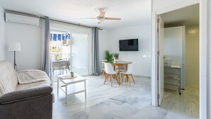 Een moderne woonkamer met een bank, een eettafel met stoelen, een plafondventilator en een aan de muur bevestigde televisie.