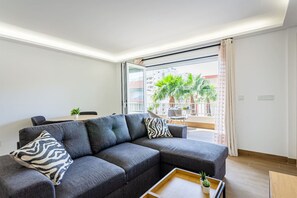 Un salon moderne avec un canapé d’angle, une table basse agrémentée d’une plante et un balcon orné de palmiers.