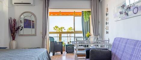 Una habitación de hotel con cama, sofá, mesa de comedor con sillas, balcón con vista al mar y un cuadro en la pared.