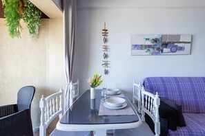 A dining area with a table set for two, a white chair, a sofa, and a wall-mounted art piece. - Mar y calma Chinasol Almuñecar (Almuñécar)