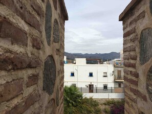 Une vue à travers une ruelle étroite bordée d’immeubles en pierre, qui mène à un bâtiment blanc doté de nombreuses fenêtres et de balcons, le tout se détachant sur un paysage montagneux.