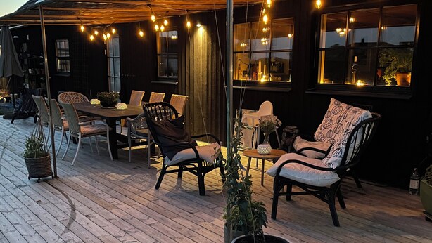 Una terraza de madera con mobiliario exterior, un área techada y luces colgantes.