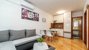 Een moderne woonkamer met een grijze bank, een eettafel met stoelen, een kleine keukenhoek en een houten vloer.