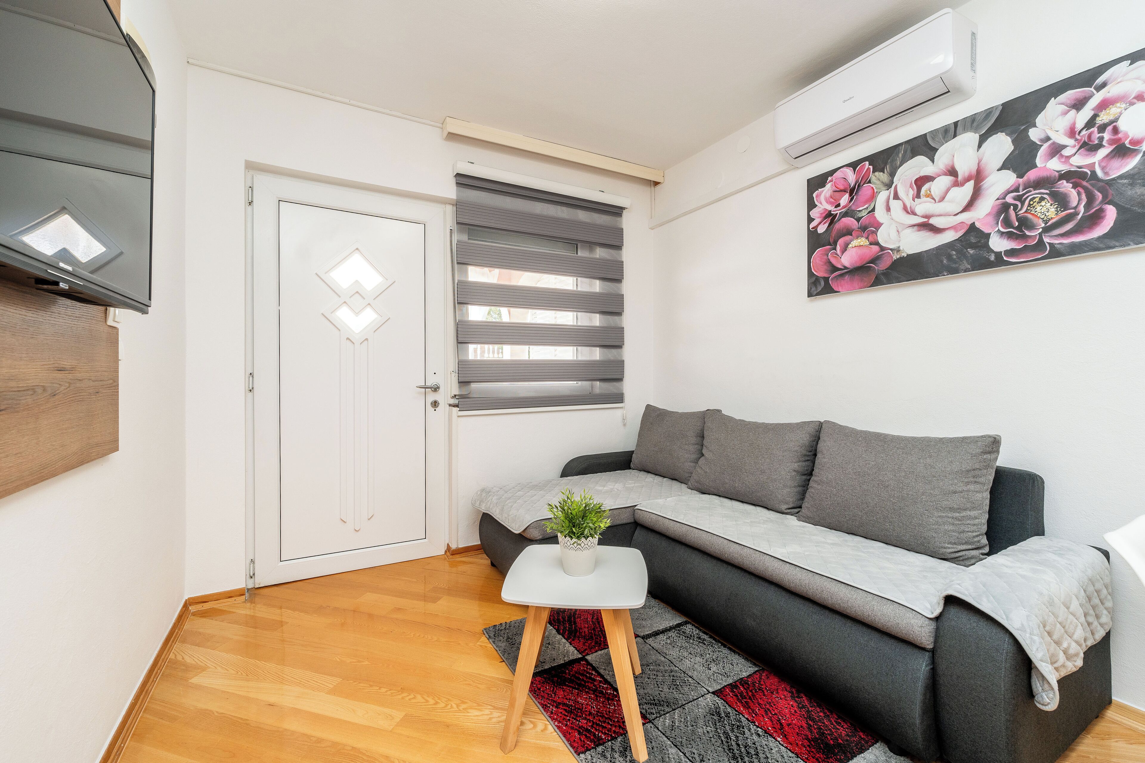 A modern living room with a grey sofa, a small white table with a plant, a red and black rug, and a large floral painting on the wall.