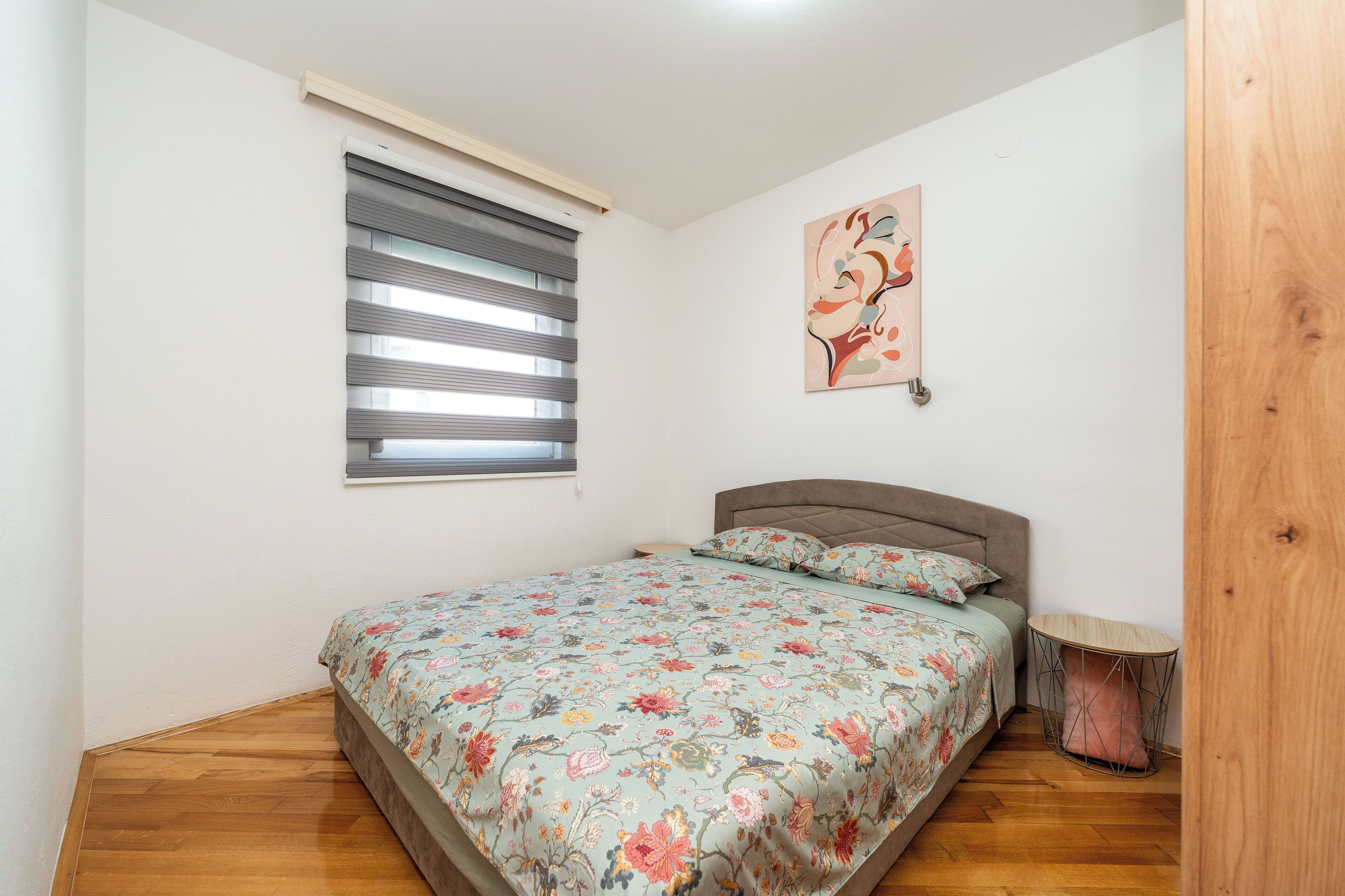 A bedroom with a bed, a window with blinds, a wall-mounted artwork, and a small wooden stool.