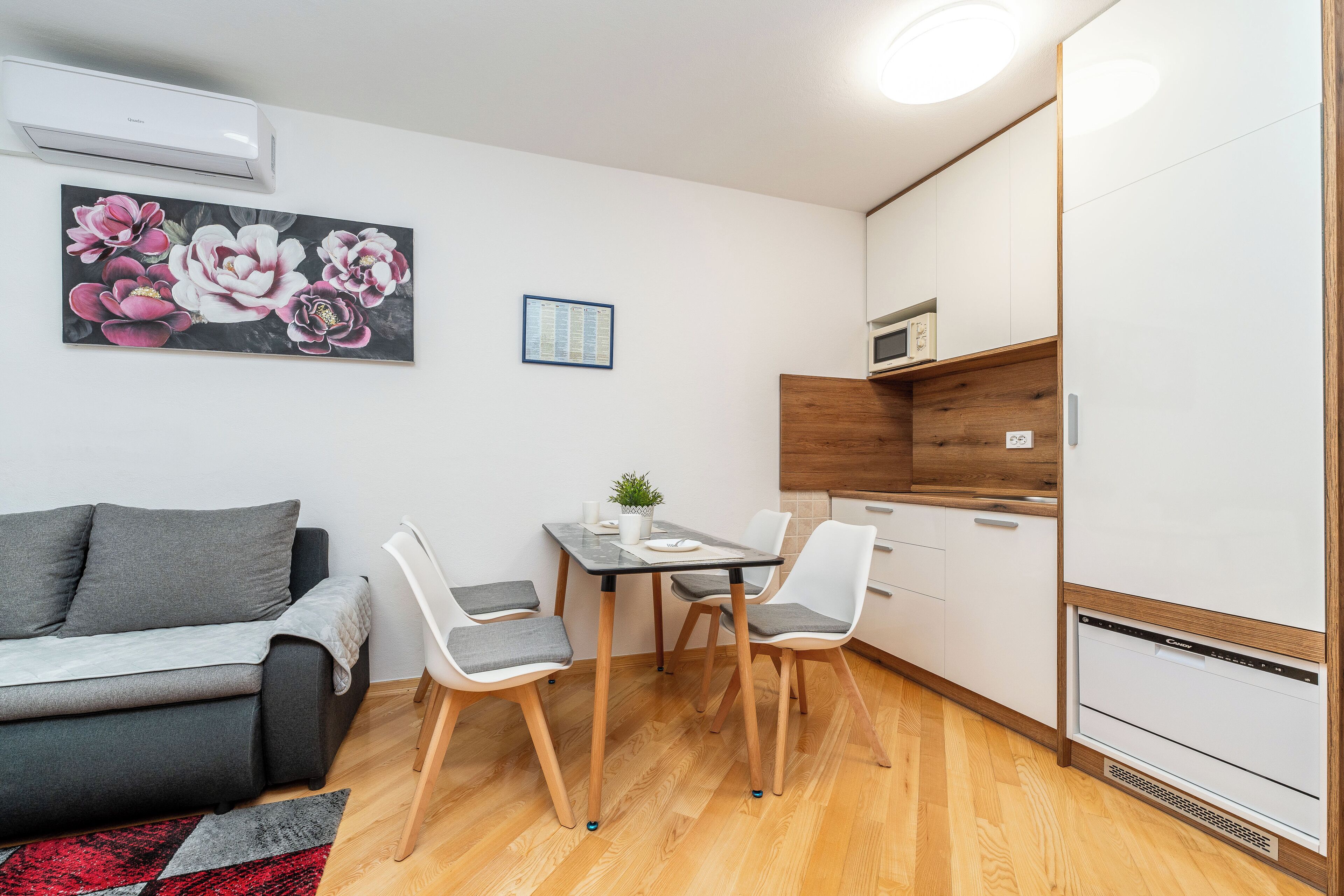 A compact living space with a sofa, dining table, chairs, a small kitchen area, and a wall-mounted air conditioner.