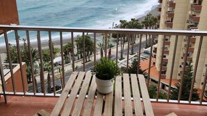 Ein Balkon mit Holztisch und -stühlen, der Blick auf Strand und Meer freigibt.