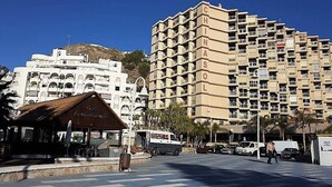A multi-story building with balconies, a street with cars, and a small building with a sign. - Nice seafront apartment (Almuñécar)