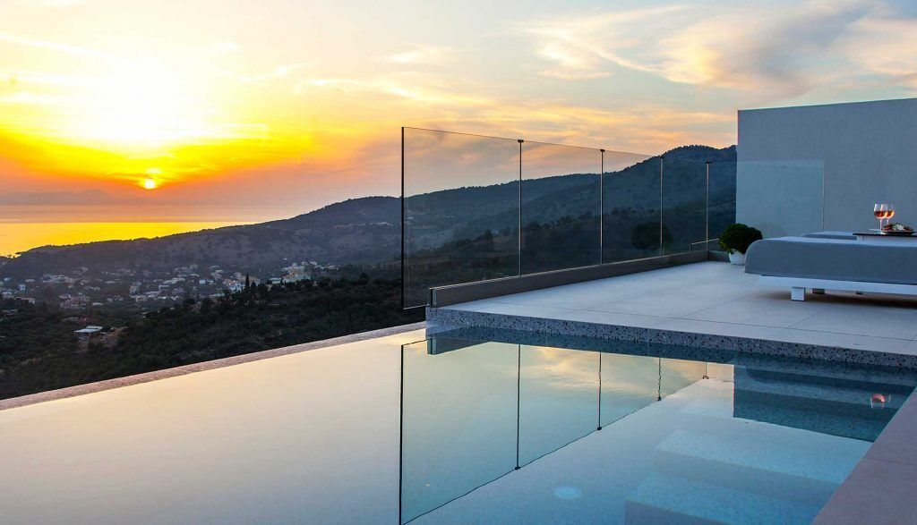 Uma piscina infinita moderna com barreira de vidro, área de estar minimalista e vista panorâmica para o pôr do sol.