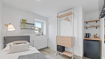 A modern bedroom with a bed, bedside lamp, dresser, mirror, and a small kitchenette with a refrigerator.
