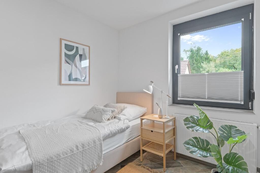 Ein Schlafzimmer mit einem Einzelbett, einem Nachttisch mit Lampe, einem Fenster mit Jalousien und einem gerahmten Bild an der Wand.