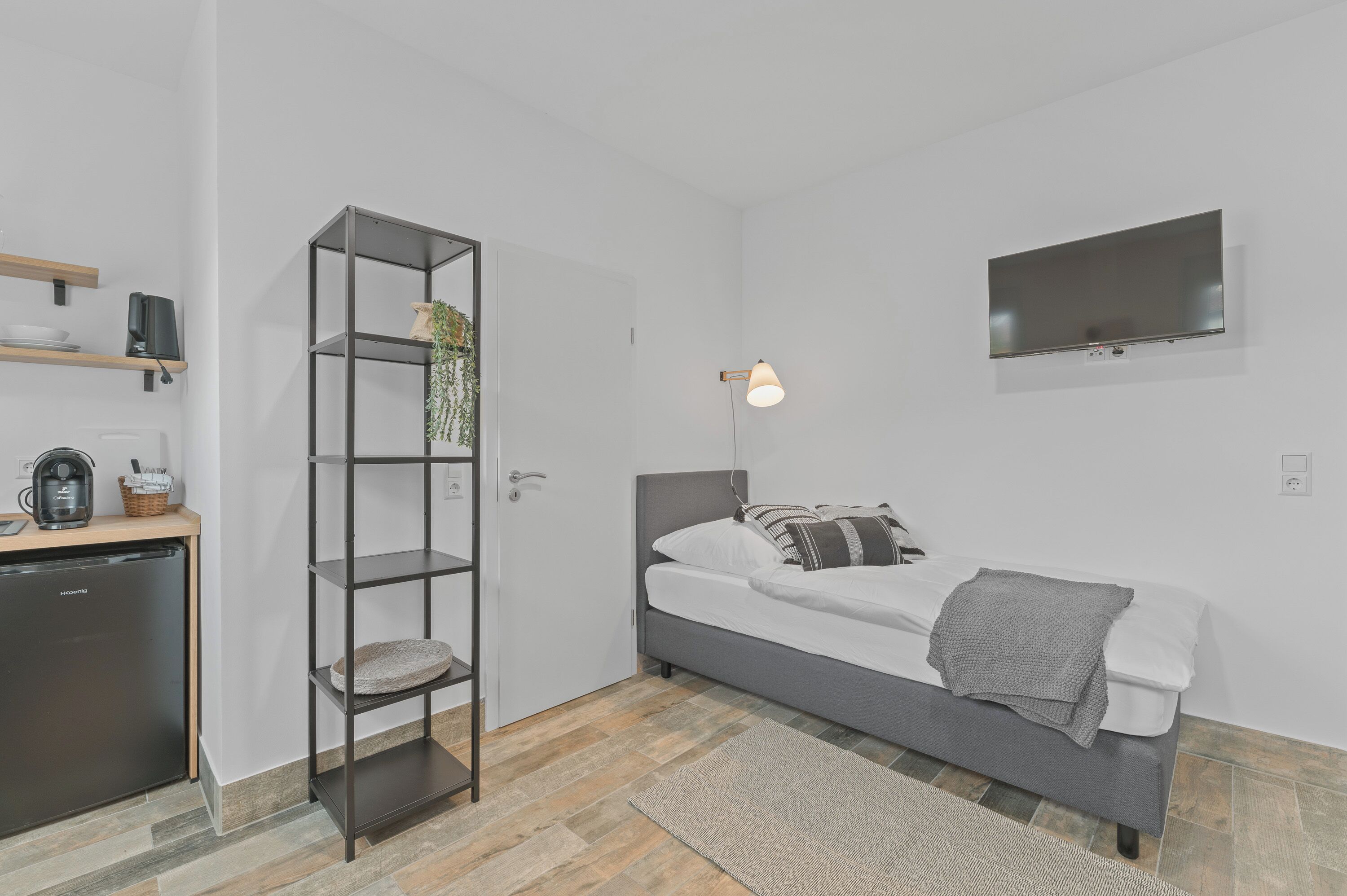A compact room with a bed, a desk with a coffee machine, a TV mounted on the wall, and a shelving unit with a plant.