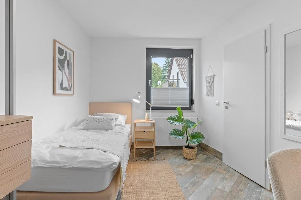 A bedroom with a bed, bedside table, lamp, potted plant, and a window with a view of a house.