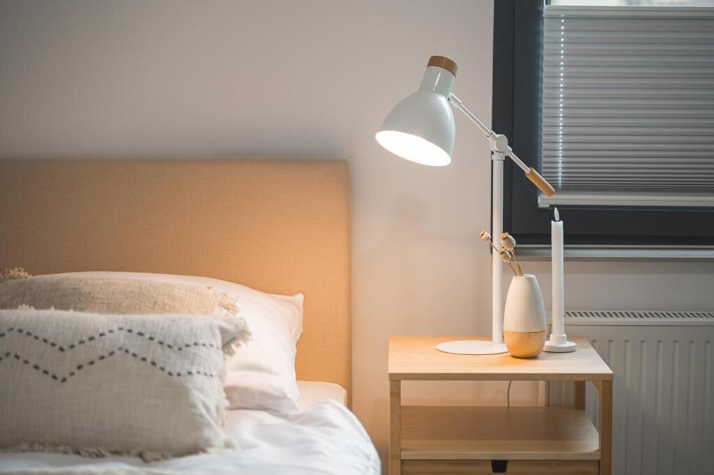 Un lit avec une tête de lit, une table de chevet avec une lampe et une fenêtre avec des stores.