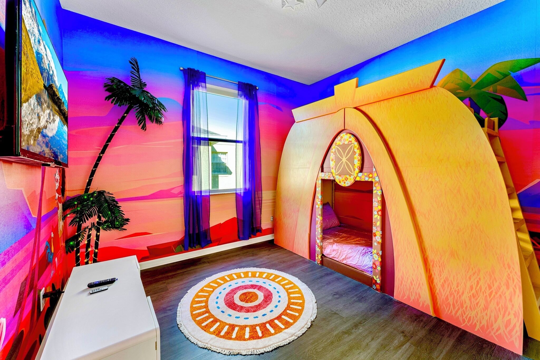 A room with a bed, a TV, a window with curtains, a small table, and a colorful wall.