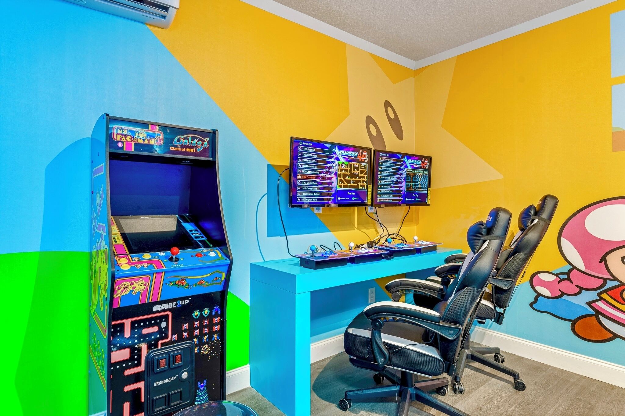 A gaming room with two arcade machines, a desk with monitors, and gaming chairs.