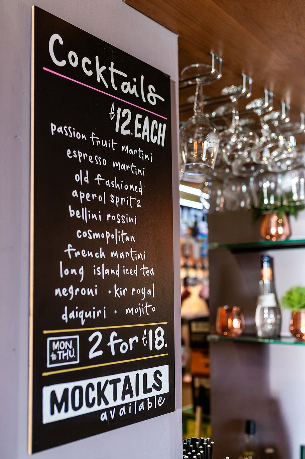 En drinklista med olika sorters drinkar och priserna, en skylt som visar att alkoholfria drinkar finns tillgängliga, och en bakgrund med upphängda glas och flaskor.