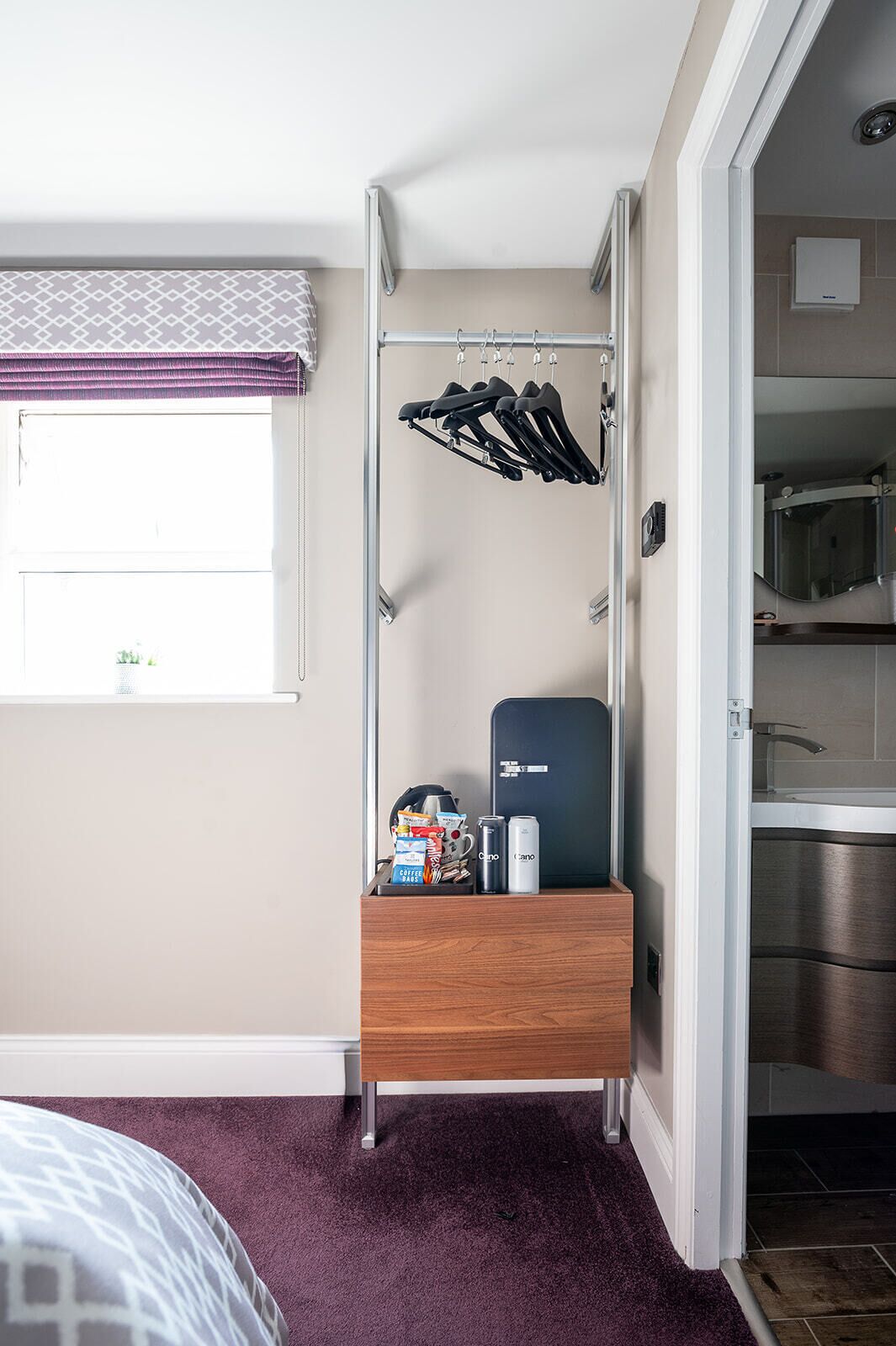 A hotel room with a built-in closet, a small wooden dresser, a window with a patterned curtain, and a door leading to a kitchenette.
