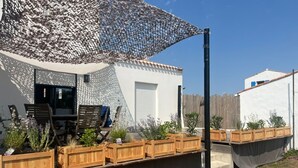A patio area with a table and chairs, wooden planters with plants, and a large umbrella. - Cozy house near the beach - 7 guests (La Guérinière)