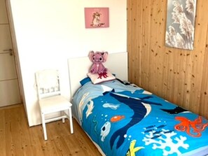 Une chambre à coucher avec un lit recouvert d’une courtepointe aux motifs bleus évoquant l’océan, une chaise blanche, un jouet en peluche rose et un tableau encadré accroché au mur.