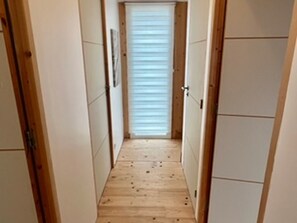 Ein schmaler Korridor mit Holzdielen, weißen Wänden und einem Fenster mit Jalousien.