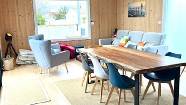 A room with a wooden dining table, blue chairs, a grey sofa, and a window with a view of greenery. - Cozy house near the beach - 7 guests (La Guérinière)
